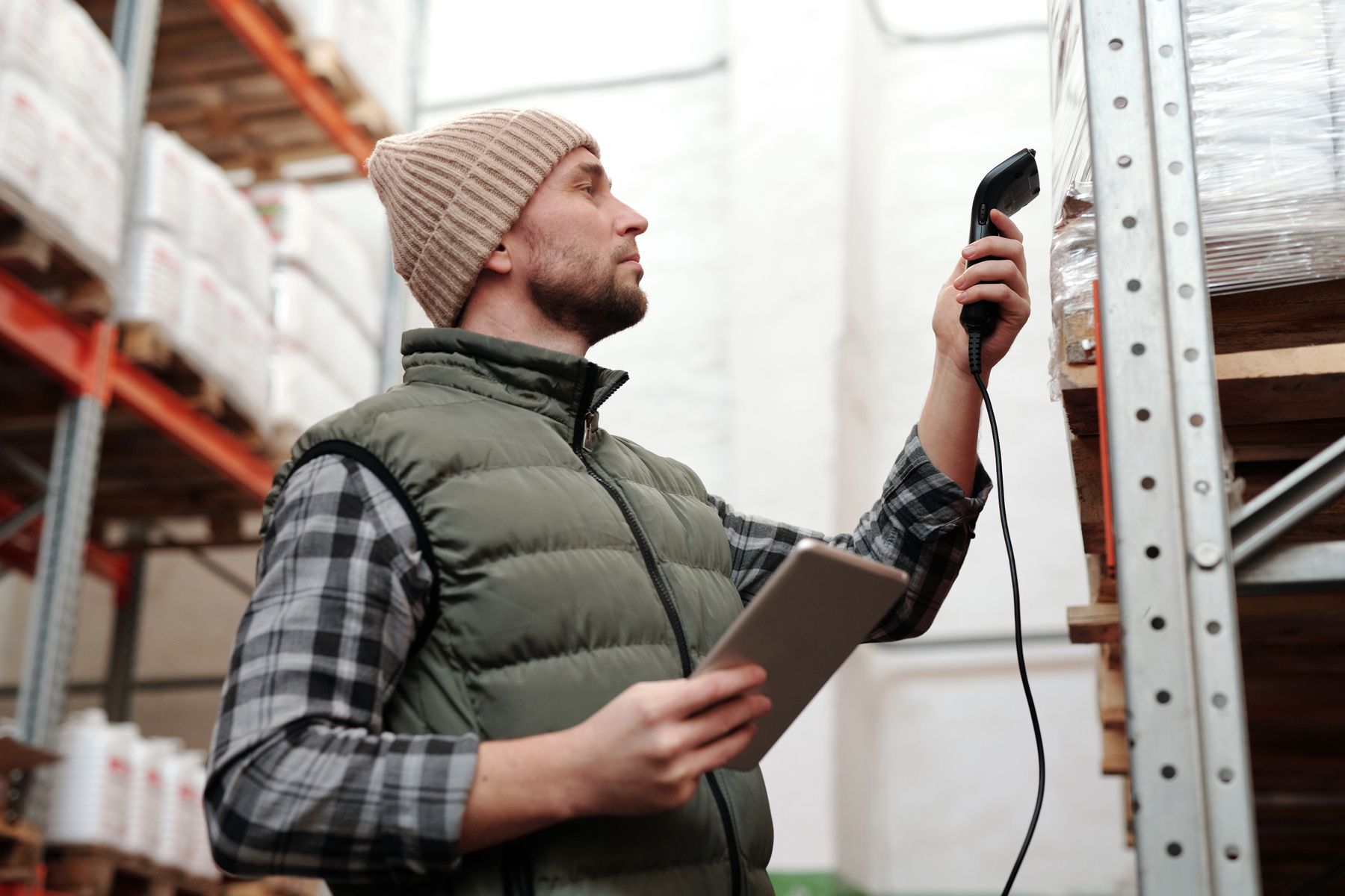 Magasinier en gilet sans manches qui scanne un colis en rayonnage, une tablette à la main